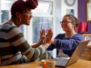 support-worker-and-young-girl-living-with-down-syndrome-playing-cheerfully-at-home-in-front-of-computer-clapping-hands-scaled-1 (1)