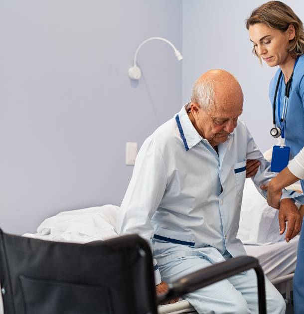 Lovely nurse assisting senior man to get up from bed at hospital. Caring nurse supporting elderly patient while getting up from bed and move towards wheelchair at nursing home. Disabled man standing up at care centre while caregiver helping him to get up in the morning.